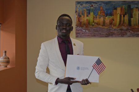 Oballa standing and posing with his citizenship certifiacte envelope and a small USA flag. There is a sculpture piece on the wall behind him of the Twin Cities skyline.