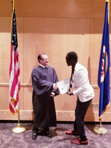 Oballa is shaking the hand of the Judge. There is a USA flag and a Minnesota flag and the wall behind them is wooden.