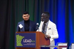 Oballa in a white suit jacket speaking at a LeadMN podium. The podium has a LeadMN sign on it. Another man stands next to Oballa and behind them there is a deep blue curtain.